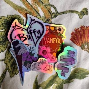 May include: A set of five iridescent stickers featuring the words "Buffy" and "Vampyr", a floral design, a hand holding a stake, a heart with a Celtic knot design, and a banner that reads "She saved the world a lot".