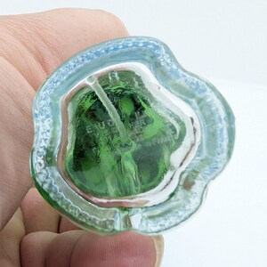 May include: A clear glass perfume bottle with a green interior. The bottle has a unique, irregular shape with a light blue rim. The text "E. VOUCHER" is visible inside the bottle. The bottle is held in a hand.