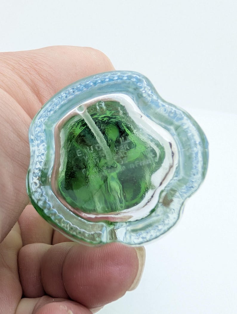 May include: A clear glass perfume bottle with a green interior. The bottle has a unique, irregular shape with a light blue rim. The text "E. VOUCHER" is visible inside the bottle. The bottle is held in a hand.