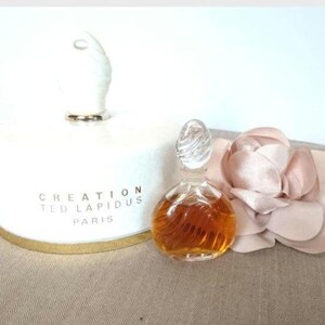 Könnte beinhalten: Eine weiße Keramikflasche mit goldfarbenem Rand und dem Text "CREATION TED LAPIDUS PARIS" auf der Vorderseite. Eine kleine, durchsichtige Glasflasche mit Parfüm und brauner Flüssigkeit steht neben der Keramikflasche.