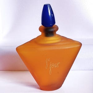 以下が含まれることがあります： 青いプラスチック製のキャップが付いた、フロスト加工された琥珀色のガラス製の香水瓶。瓶は三角形で、金色の文字で「Le Jour」と書かれています。