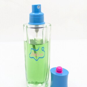 May include: A clear glass perfume bottle with a blue sprayer and cap. The bottle contains a green liquid and has the word "Salsa" in pink script. The cap is blue with a pink top.