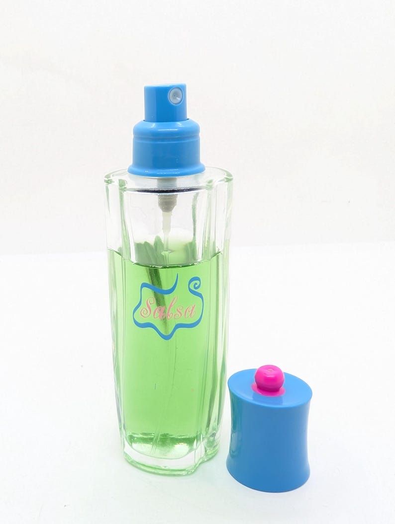 May include: A clear glass perfume bottle with a blue sprayer and cap. The bottle contains a green liquid and has the word "Salsa" in pink script. The cap is blue with a pink top.