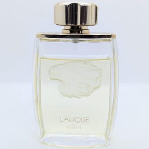 Könnte beinhalten: Ein klarer Glasparfümflakon mit goldener Kappe und Zierleiste. Die Flasche enthält eine hellgelbe Flüssigkeit und weist ein erhabenes Löwenkopfdesign auf. Der Text "LALIQUE POUR HOMME" ist auf der Vorderseite aufgedruckt.