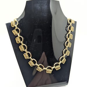 May include: A gold-toned necklace with a unique design, displayed on a black mannequin. The necklace features a series of interconnected links, each adorned with a coiled, spiral-like element. The jewelry is set against a white background.