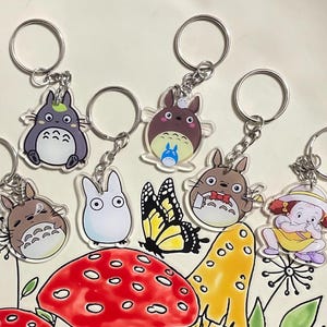 Może przedstawiać: Zestaw 7 kolorowych breloków z postaciami z kreskówki z filmu animowanego Mój sąsiad Totoro. Breloki są wykonane z przezroczystego akrylu i przedstawiają różne postacie z filmu, w tym Totoro, Kota-busa i małą dziewczynkę.