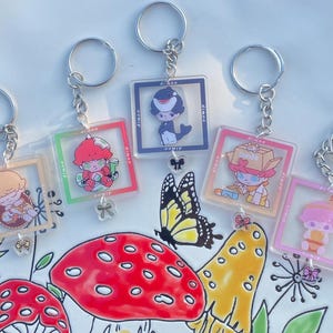 May include: Five colorful keychains with different cartoon characters on them. Each keychain has a silver metal ring and a clear acrylic charm with a cartoon character inside. The charms are decorated with colorful details and glitter. The keychains are arranged on a white surface with a floral pattern.