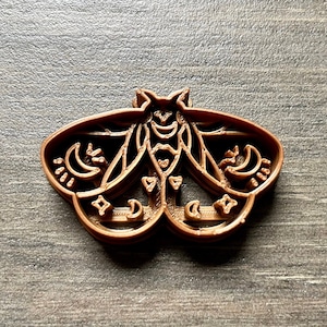 May include: A brown plastic cookie cutter shaped like a moth with intricate details, including crescent moons and stars.