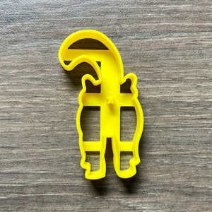 May include: Yellow plastic cookie cutter shaped like a cartoon raccoon standing on its hind legs with its tail curled over its head.