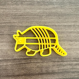May include: A bright yellow armadillo-shaped cookie cutter. The cutter is made of plastic and has a detailed outline of the animal, including its segmented shell and tail. The cutter is designed for baking and crafting.