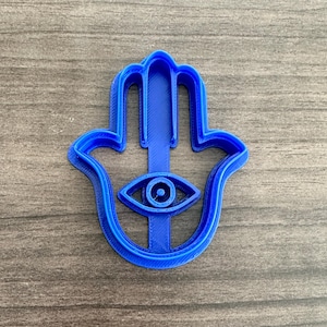 May include: A blue plastic cookie cutter in the shape of a Hamsa hand with an eye in the center. The cutter is a solid color and sits on a wooden surface.