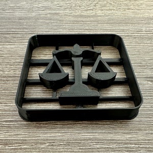 May include: Black plastic cookie cutter shaped like a scale of justice. The cutter is in focus and the background is a wood grain surface.