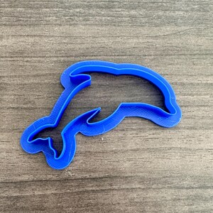 May include: A bright blue dolphin-shaped cookie cutter. The cutter is made of plastic and has a smooth, clean edge. The dolphin is depicted in a swimming pose, with its tail curved and its mouth slightly open. The cutter is designed for use in baking.