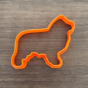May include: An orange plastic cookie cutter in the shape of a dog. The cutter is a solid outline of the dog's body, with details for the legs and head. The cutter is on a wooden surface.