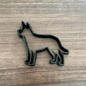 May include: A black plastic cookie cutter in the shape of a German Shepherd dog. The cutter is a silhouette of the dog, with detailed legs, tail, and head. The cutter is placed on a wooden surface.