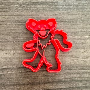 May include: A red plastic cookie cutter in the shape of a dancing bear. The bear has a smiling face, a jagged chest, and outstretched arms and legs. The cutter is on a wooden surface.