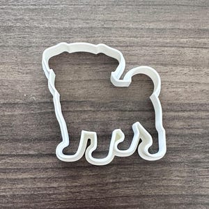 May include: A white plastic cookie cutter in the shape of a pug dog. The cutter has a simple outline with details for the head, body, legs, and tail. The cutter is designed for making pug-shaped cookies or other treats.