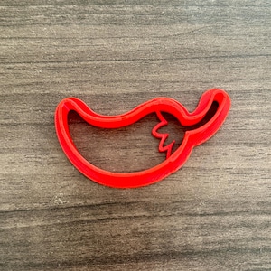 May include: A bright red plastic chili pepper-shaped cookie cutter. The cutter is a single piece with a smooth, rounded outline and a detailed stem. The cutter is designed for baking and crafting.
