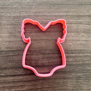 May include: A pink plastic onesie cookie cutter. The cutter is in the shape of a baby bodysuit with short sleeves. The cutter is made of pink plastic and is designed for use in baking or crafting projects.