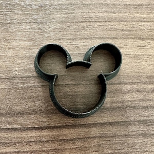 May include: A black cookie cutter in the shape of Mickey Mouse's head. The cutter is made of a dark material and has a textured surface. The cutter is placed on a wooden surface.