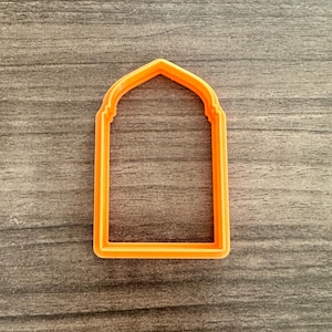 May include: An orange plastic cookie cutter in the shape of an arched window or doorway. The cutter is made of a single piece of plastic and has a smooth, clean design. The cutter is set against a gray wooden surface.