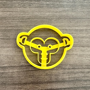 May include: A yellow plastic monkey-shaped cookie cutter. The cutter features a circular outline with monkey face details, including ears, eyes, and a mouth. The cutter is designed for baking and crafting.