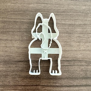May include: A white plastic cookie cutter in the shape of a dog's rear end, including the tail, legs, and ears. The cutter is designed to create dog-shaped cookies or treats.
