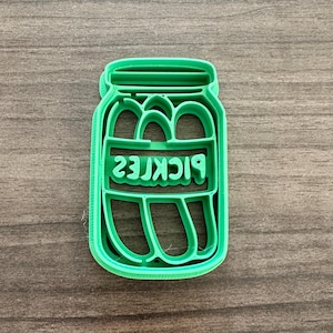 May include: A bright green plastic cookie cutter in the shape of a pickle jar. The word "PICKLES" is printed in reverse inside the jar. The cutter is sitting on a wooden surface.