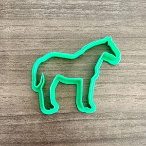 May include: A bright green horse-shaped cookie cutter. The cutter is made of plastic and has a simple outline of a horse. The cutter is on a wooden surface.