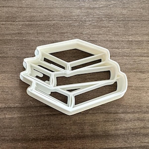 May include: A white plastic cookie cutter in the shape of a stack of books. The cutter has a detailed outline of the books, with visible pages and covers. The cutter is designed for baking and crafting.
