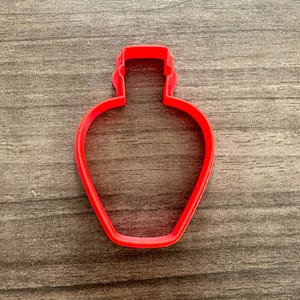 May include: A bright red, plastic cookie cutter in the shape of a perfume bottle. The cutter is made of a single piece of plastic and has a simple, clean design. The cutter is sitting on a wooden surface.