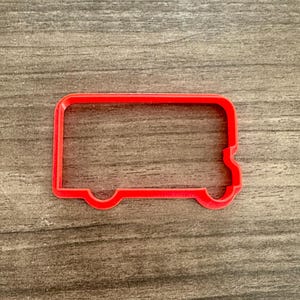 May include: A red plastic cookie cutter in the shape of a school bus. The cutter has a rectangular body with rounded corners and two small wheel cutouts. The cutter is on a wooden surface.