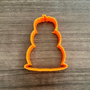 May include: An orange plastic cookie cutter in the shape of a snowman. The cutter has a three-tiered body with a small, rounded head and a flat base. The cutter is made of a bright orange plastic.