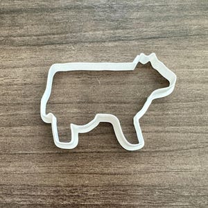 May include: A white plastic cookie cutter in the shape of a cow. The cutter has a simple outline with the cow's body, legs, and head clearly defined. The cutter is placed on a wooden surface.
