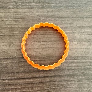 May include: An orange, circular cookie cutter with a scalloped edge. The cutter is made of plastic and is set against a dark wood grain background. The cutter is a baking tool for making cookies.