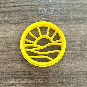 May include: A bright yellow, circular cookie cutter featuring a sunrise design. The cutter has a sun with rays and rolling hills. The cutter is made of plastic and is on a wooden surface.