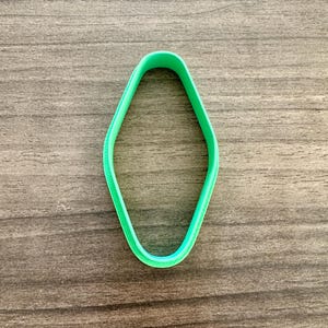 May include: A bright green, diamond-shaped cookie cutter. The cutter is made of plastic and has a smooth, rounded edge. The cutter is set against a wooden background.