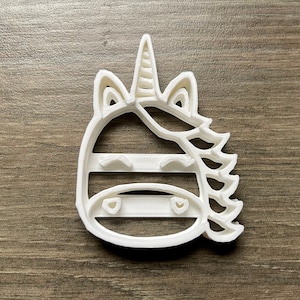 May include: White plastic cookie cutter shaped like a unicorn with a striped horn and a mane. The unicorn has closed eyes and a small mouth.