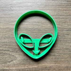 May include: A bright green alien face cookie cutter. The cutter features a circular head shape with large eyes, a nose, and a smiling mouth. The cookie cutter is made of plastic and is designed for baking.