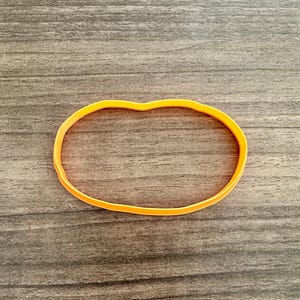May include: An orange rubber band in an oval shape, resting on a wooden surface. The band is a bright, solid color and appears to be new. The background is a dark wood grain.
