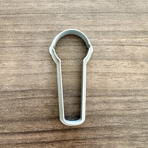 May include: A light gray plastic cookie cutter in the shape of a microphone. The cutter has a rounded top and a long, rectangular handle. The cutter is made of a smooth, solid plastic and is designed for cutting dough or fondant.