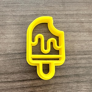 May include: A bright yellow popsicle-shaped cookie cutter. The cutter has a bite taken out of the top and a handle at the bottom. The design includes dripping details, perfect for creating fun treats.