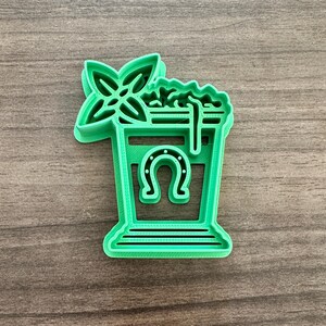 May include: A green plastic cookie cutter in the shape of a cocktail glass with a horseshoe, mint sprig, and foam detail. The cookie cutter is designed for making St. Patrick's Day themed cookies.