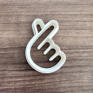 May include: A white plastic cookie cutter in the shape of a hand making a heart symbol. The cutter is on a brown wooden surface. The design features a curved index finger and thumb forming a heart.
