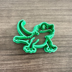 May include: A bright green plastic cookie cutter in the shape of a lizard. The lizard has a long tail, four legs, and a detailed head with an eye. The cutter is designed to create reptile-shaped cookies or treats.