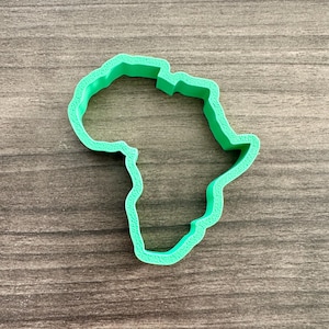 May include: A bright green cookie cutter in the shape of the African continent. The cutter is made of plastic and sits on a wooden surface. The design is a simple outline of the continent.