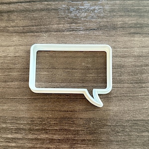 May include: A white, rectangular speech bubble cookie cutter with a rounded rectangular shape and a small tail. The cutter is made of plastic and is set against a wooden background.