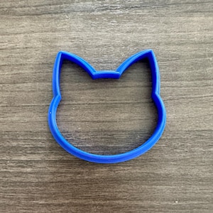 May include: A blue cat-shaped cookie cutter. The cutter is made of plastic and has a simple outline of a cat's head with pointed ears. The cutter is on a wooden surface.