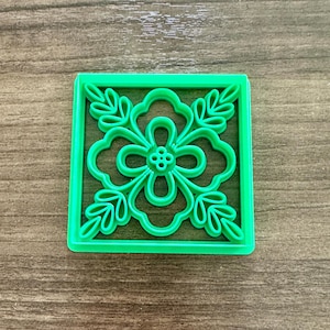 May include: A square, bright green cookie cutter featuring a floral design. The cutter has a square border with a flower and leaf pattern inside. The flower has a central cluster of small circles.