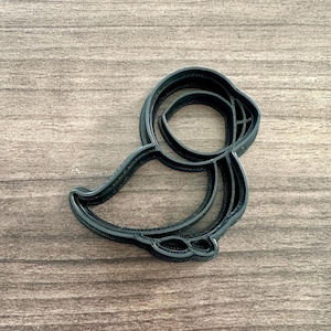 May include: A black plastic cookie cutter in the shape of a puffin bird. The cutter has a detailed outline of the bird's body, wings, and head, with a rounded shape and textured edges. The cutter is designed for baking.